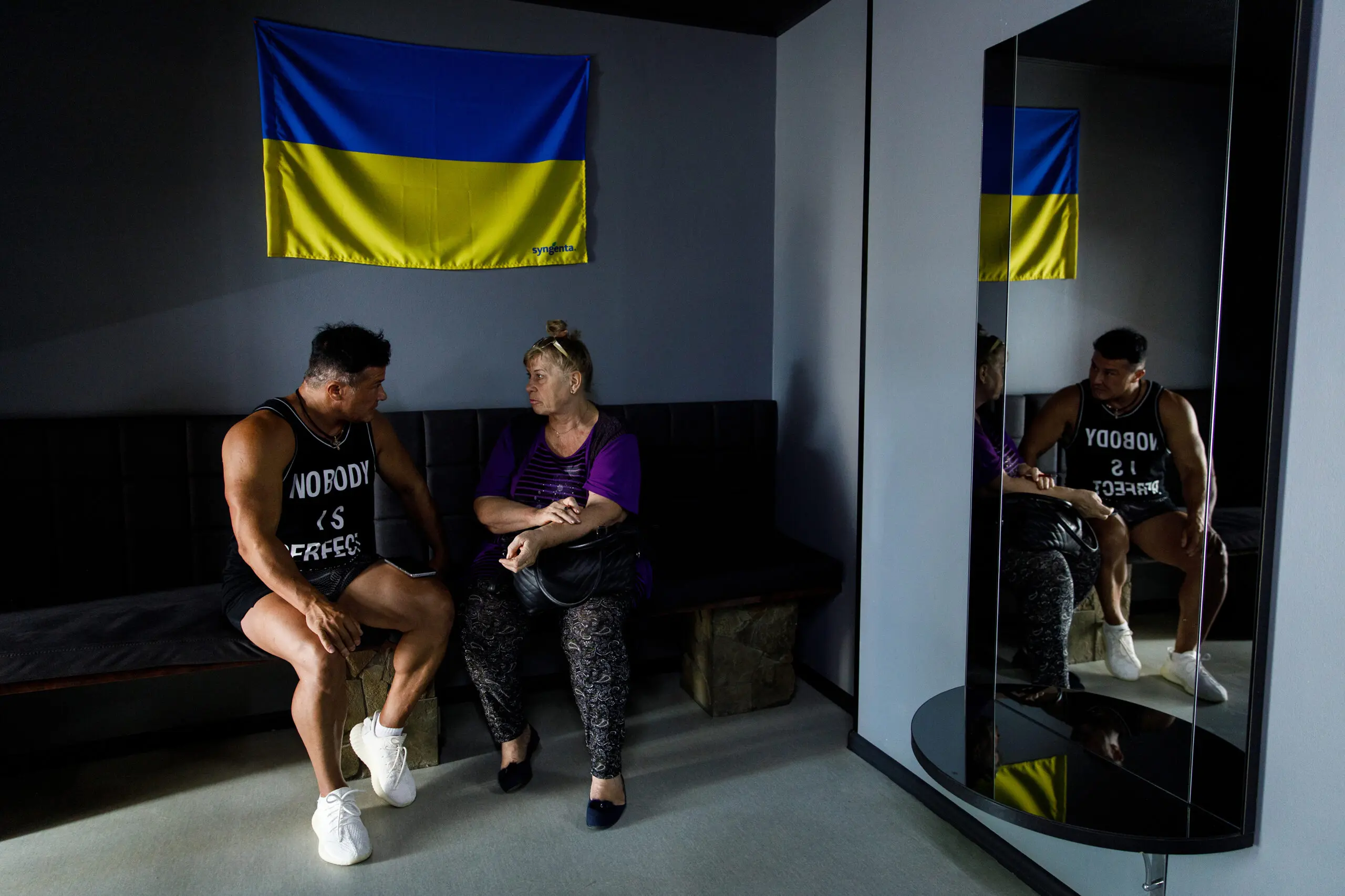 Two women sit on a couch talking, under a Ukrainian flag hanging on the wall.