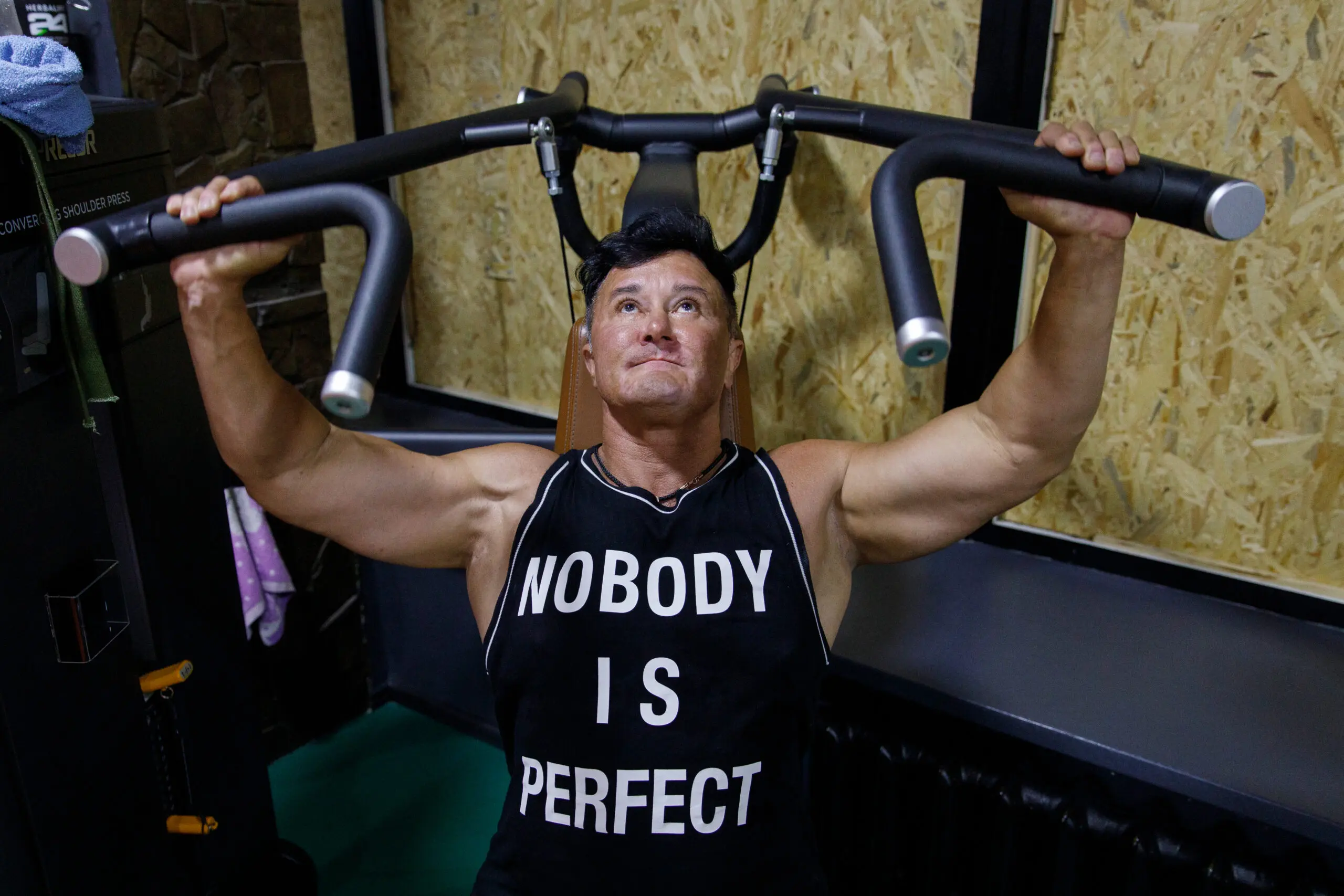 Anna Kurkurina weightlifts inside a gym, in a tank top that reads "Nobody is perfect."